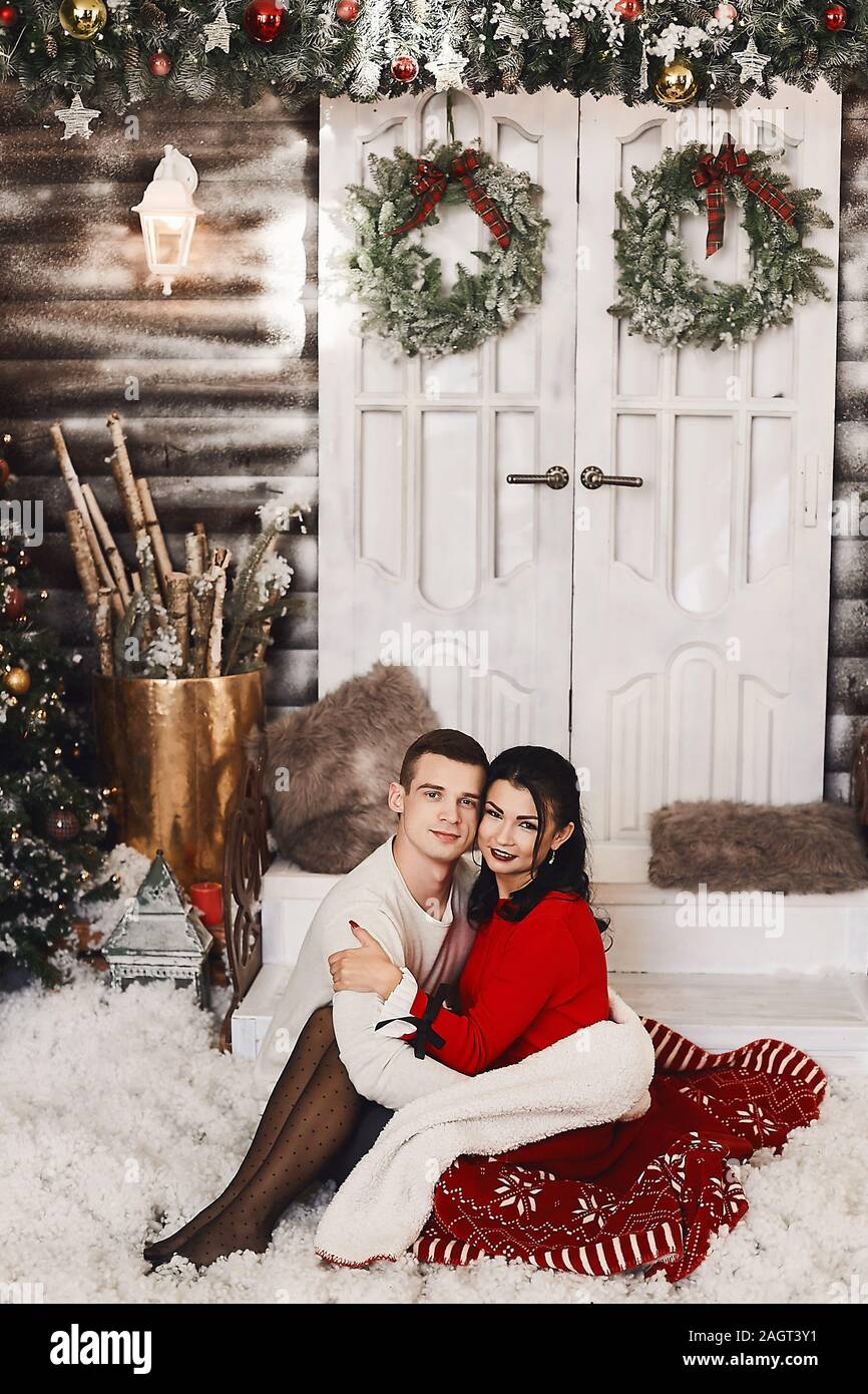 Sweet couple hugging each other on Christmas decorations background.  Handsome man and brunette young woman in red dress and stockings  celebrating Stock Photo