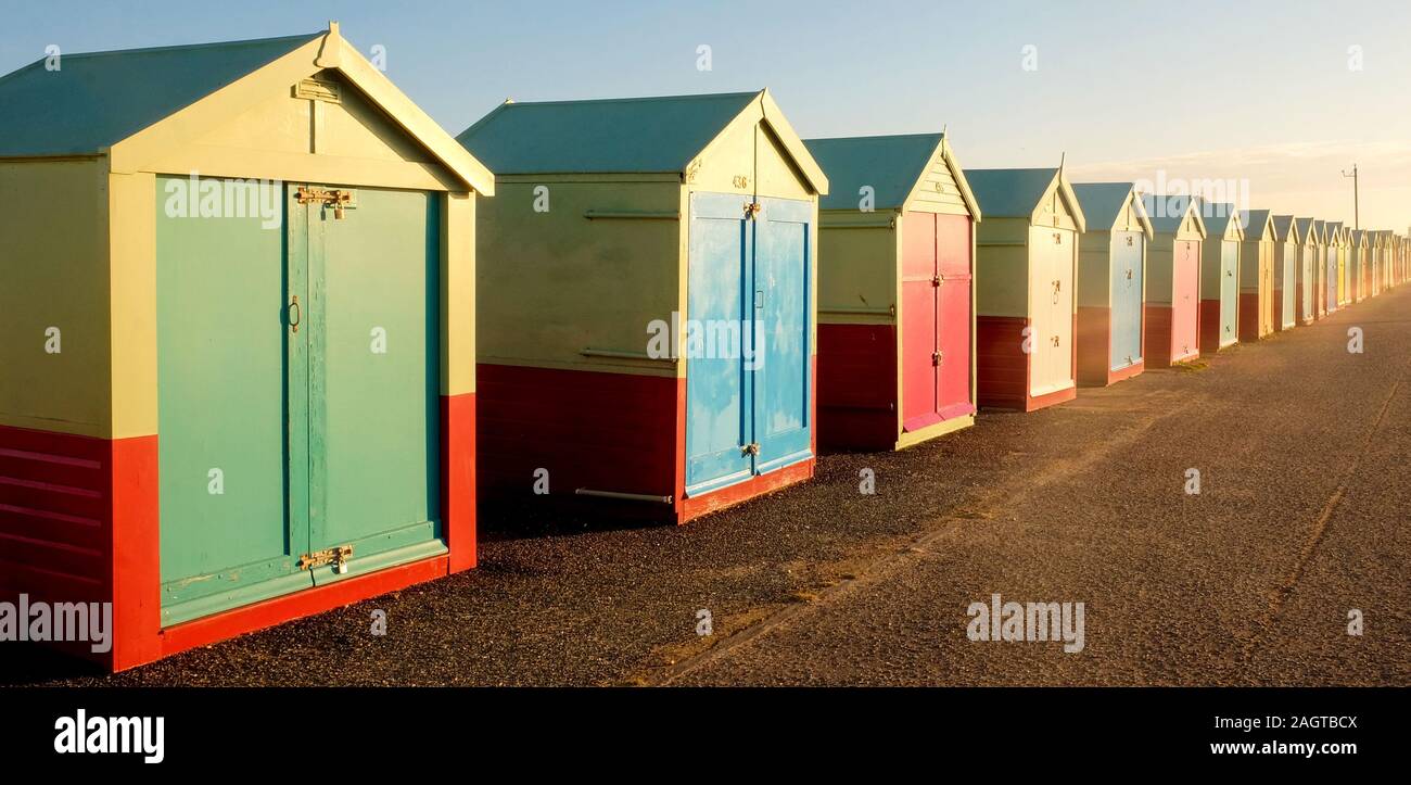 a line of 30 Beach huts with different multicoloured doors on a concrete promenade, the nearest beach huts on the left of the image are large going to Stock Photo