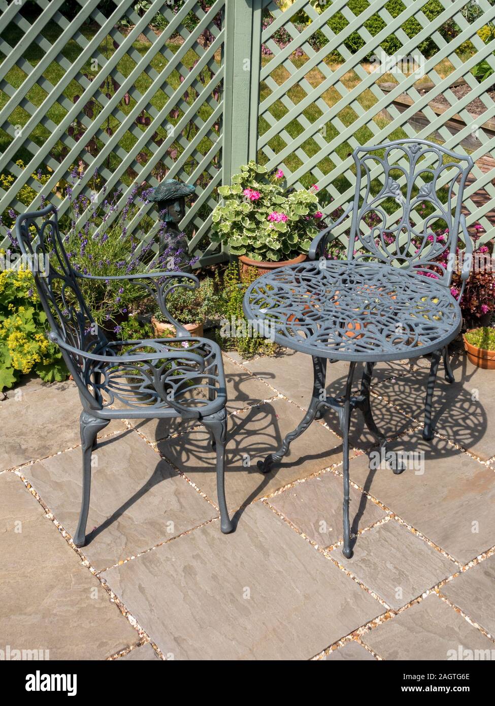 Metal table and chairs on small patio with green trellis, potted plants and natural stone slabs in English cottage garden in Summer. Stock Photo
