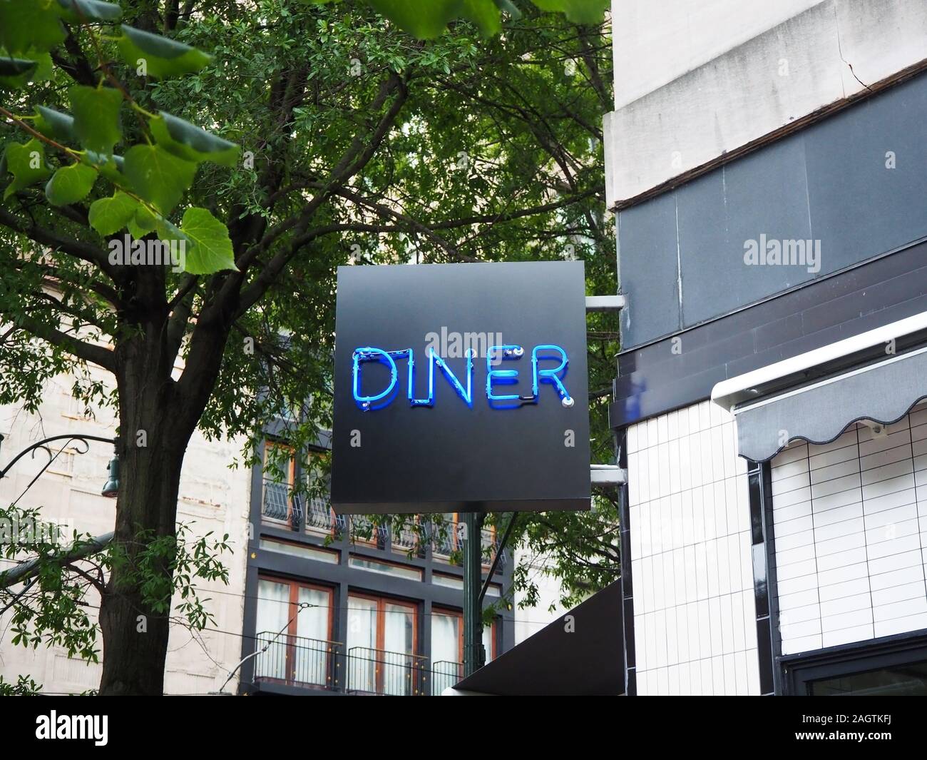 A lighted neon sign reads DINER attached to the corner of an old downtown building on a tree lined street. Stock Photo