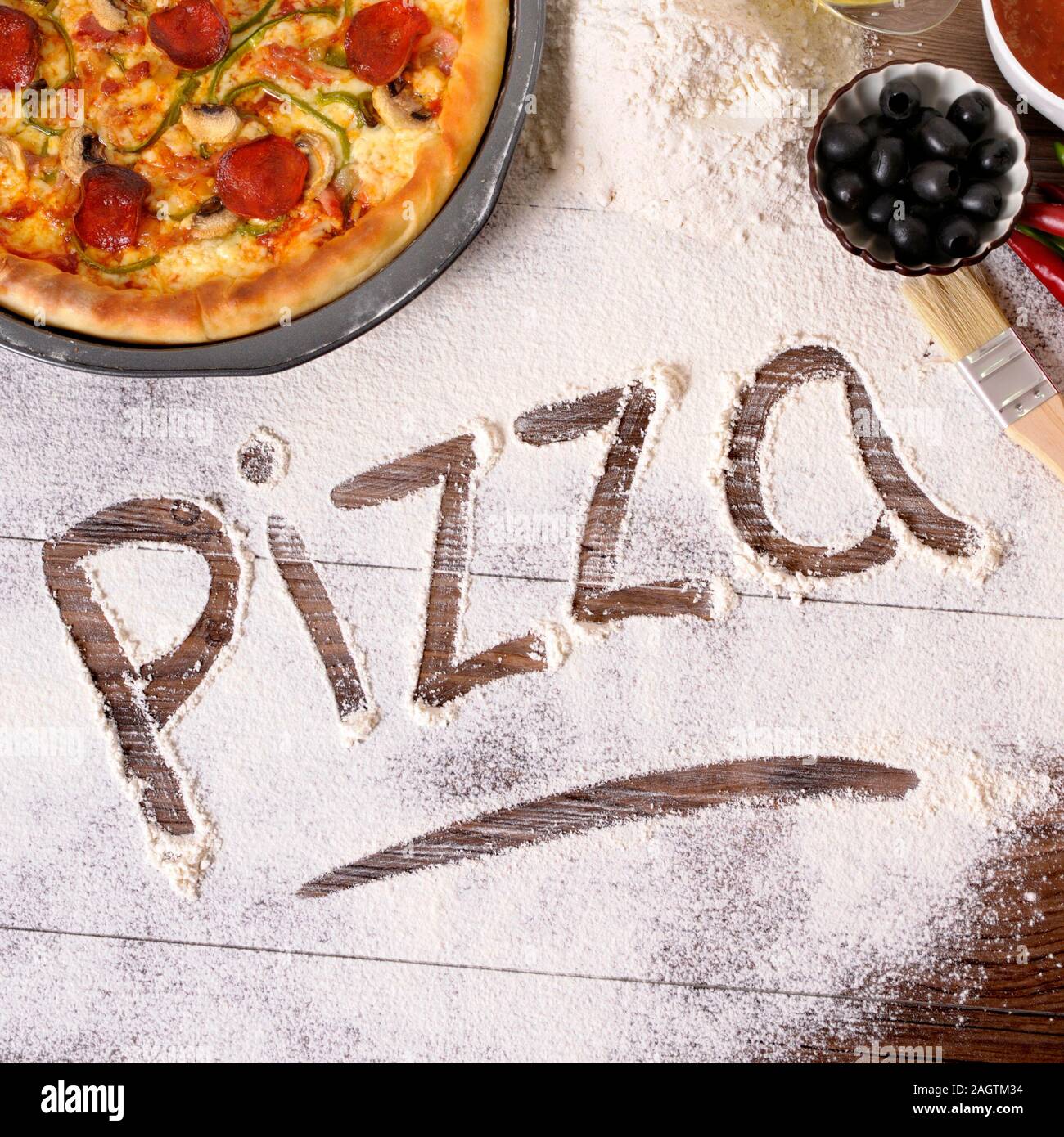 The word Pizza written in flour, with freshly baked pepperoni Pizza surrounded by various