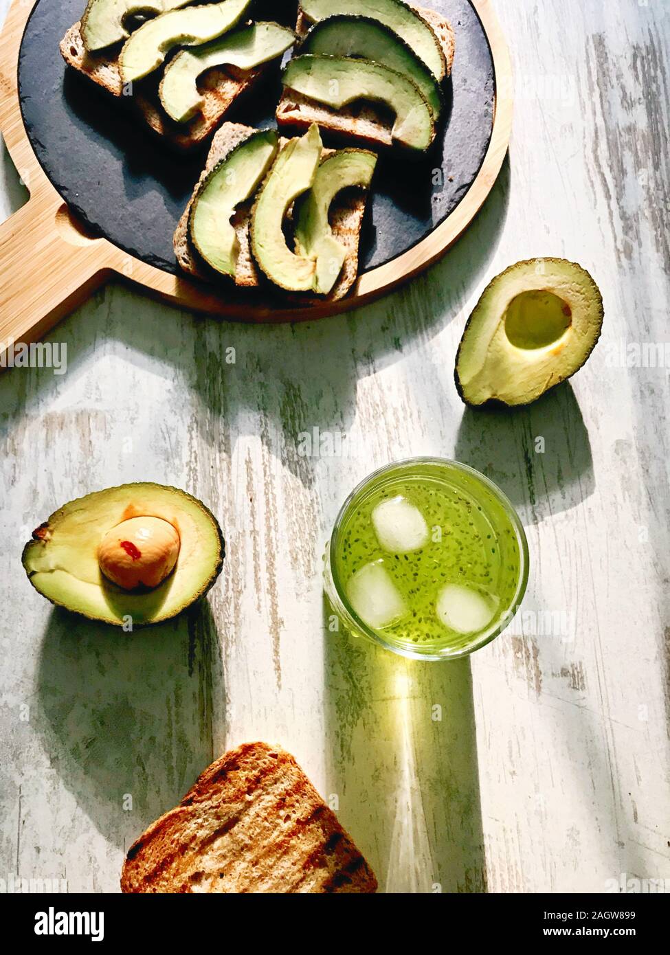 Fresh avocado on grilled toasts on round slate board with chia seeds drink and long shadow and sun shining through glass Stock Photo