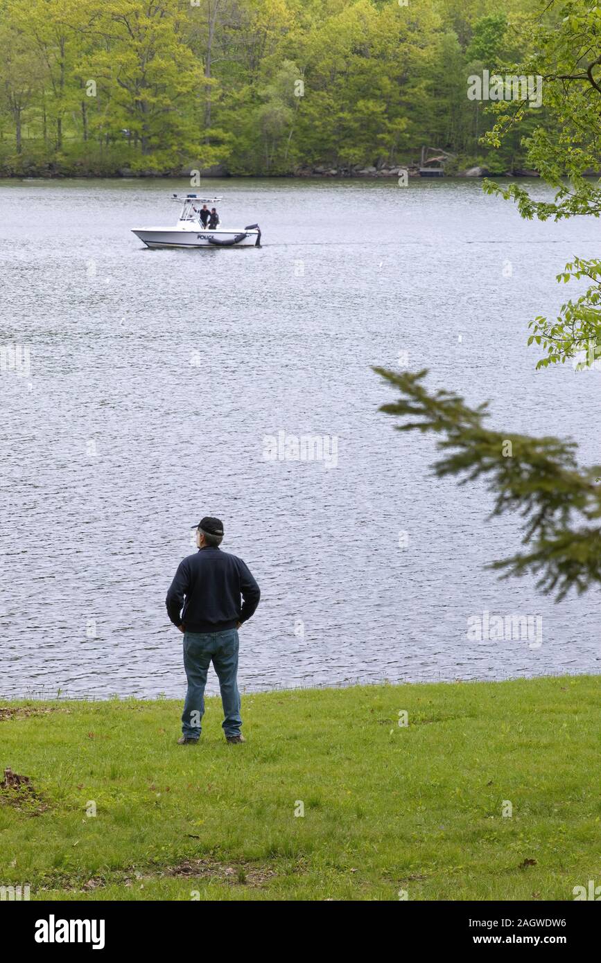 New Preston, CT USA. May 2016. Man by the shore of Lake Waramaug as a Police patrol motor boat passes by in the background. Stock Photo