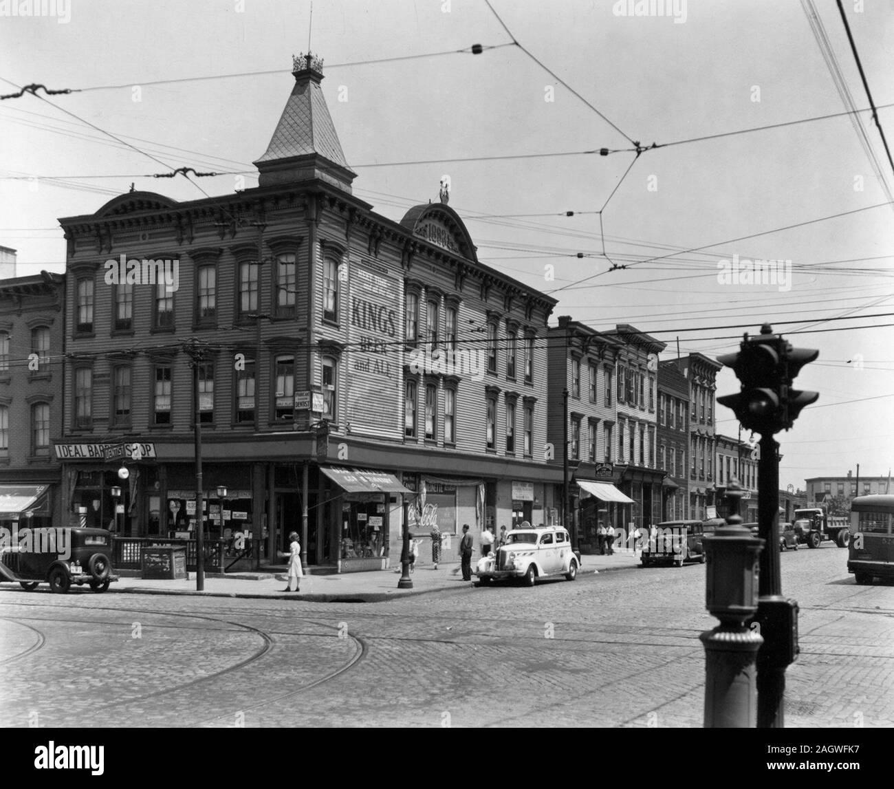 Clapboard buildings, cobblestone streets with trolley tracks, cars, including a taxi and an entrance to the subway on the corner. Graham and Metropolitan Avenues, Brooklyn. ca. 1937 Stock Photo