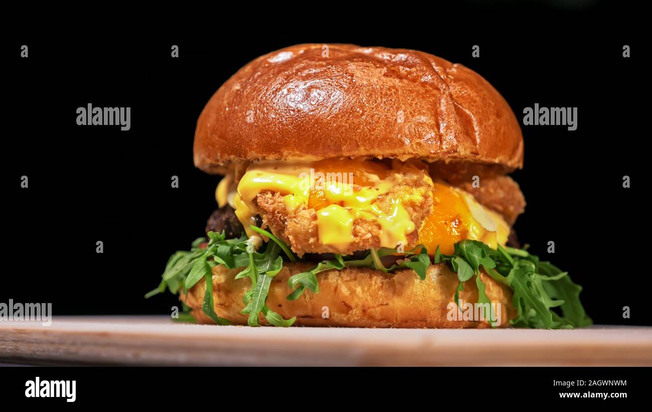 Craft burger is cooking on black background. Consist: sauce, arugula, tomato, red onion rings fries, cucumber, cheese, air bun and marble meat beef. Stock Photo