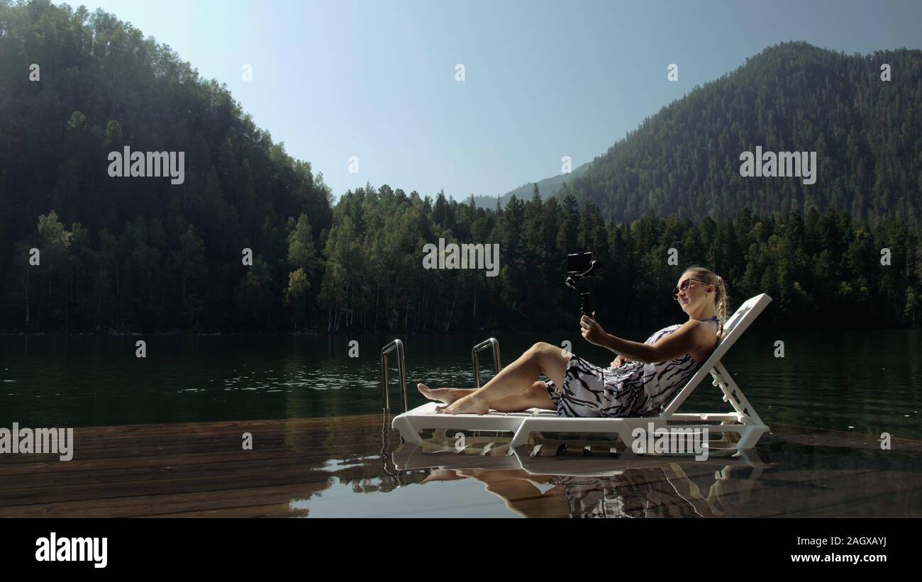 Woman shooting on handheld film gimbal stabilization for smartphone. Girl lie sunbed on pier make selfie. Blogger broadcast video vlogging. Stock Photo