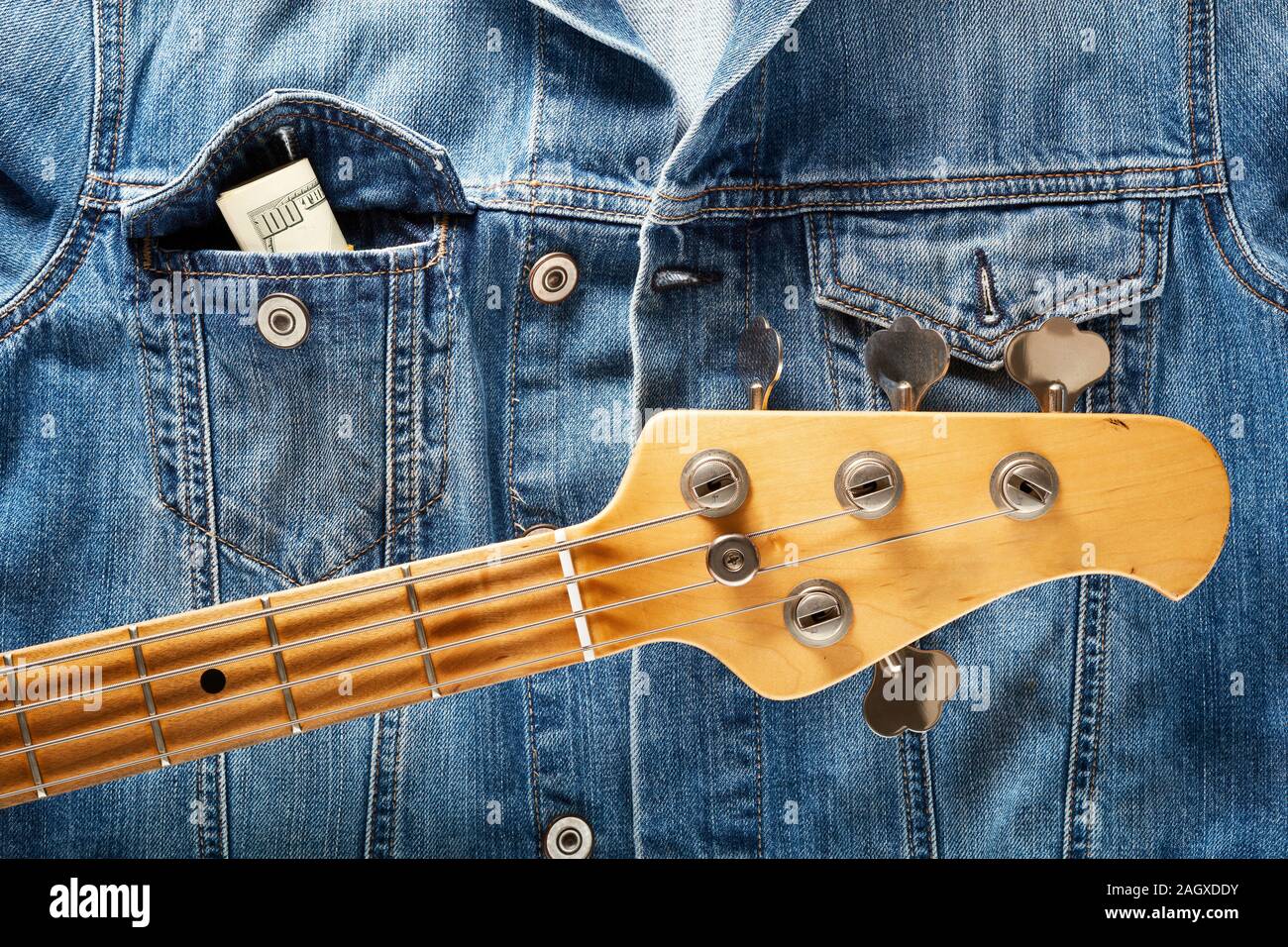 Bass guitar headstock and one dollar bills in the pocket of  denim jeans jacket. Concept of  making or earning money in music industry. Stock Photo