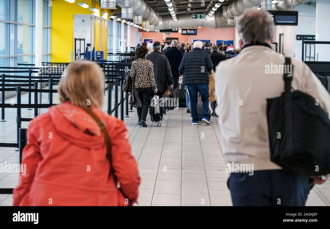 Group of anonymous people at airport gate line to board an airplane, queue crowd, pulling their trolley seen from behind. Stock Photo