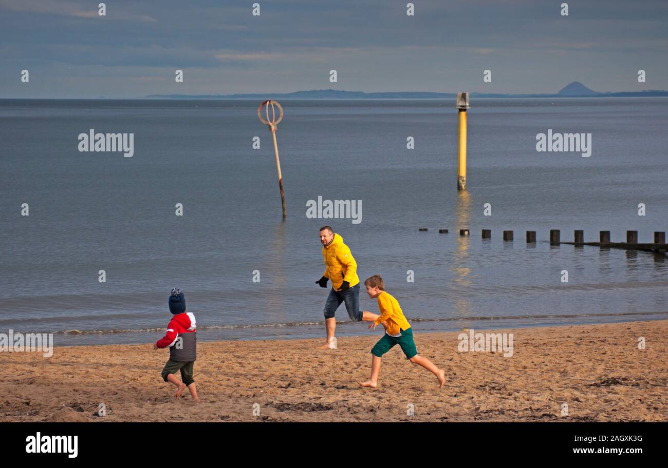 Portobello Beach, Edinburgh, Scotland, UK. 22nd December 2019. Cool temperature of 4 degrees with low sunshine on the coast encouraging people young and old to enjoy the fresh air along the sandy beach. Pictured Man with young boys playing tig while running in barefeet. Berwick Law in the background. Credit: Arch White/Alamy Live News. Stock Photo