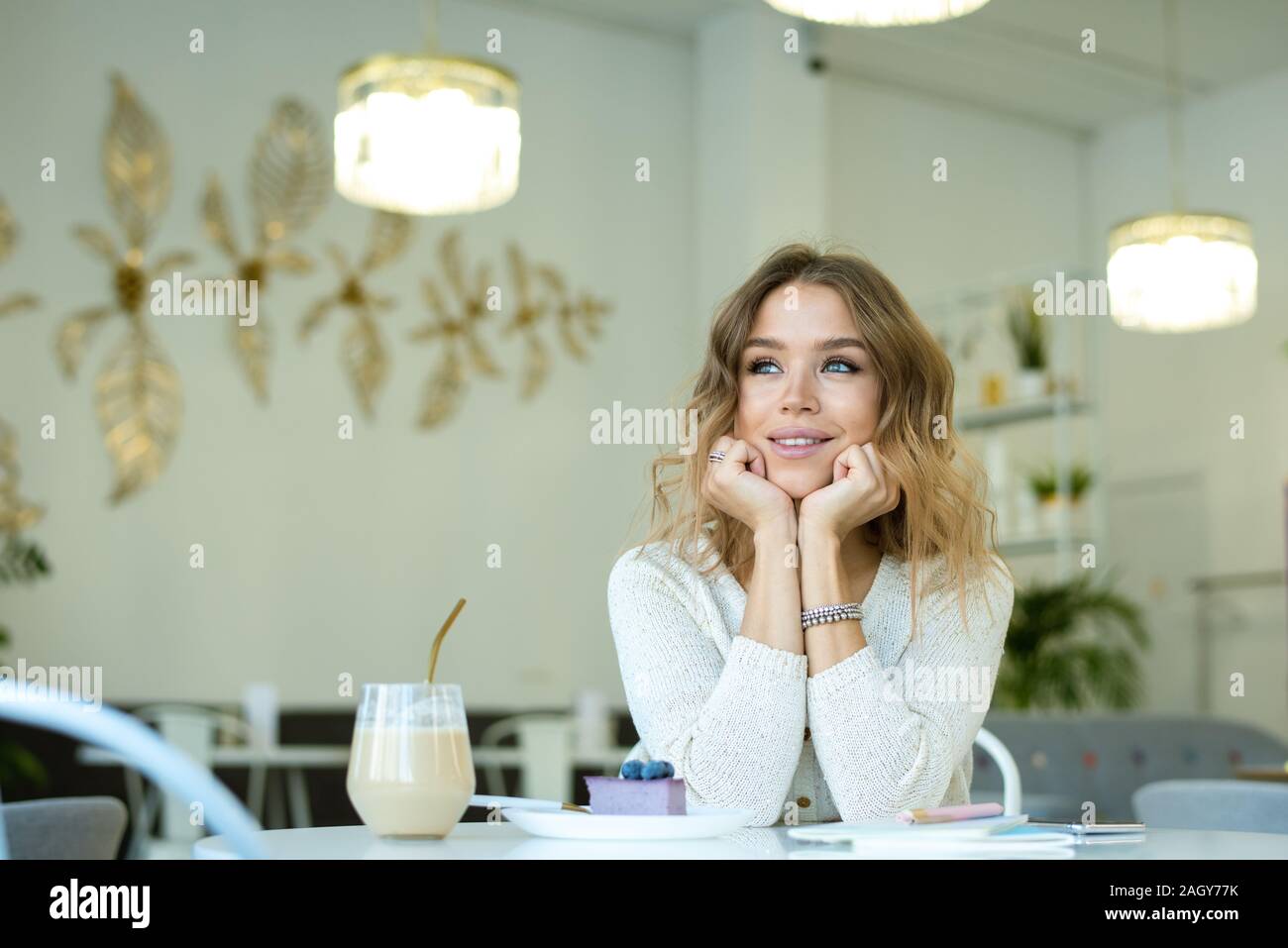 Young cheerful woman thinking of something nice while relaxing in cozy cafe by table and having cappuccino and dessert Stock Photo