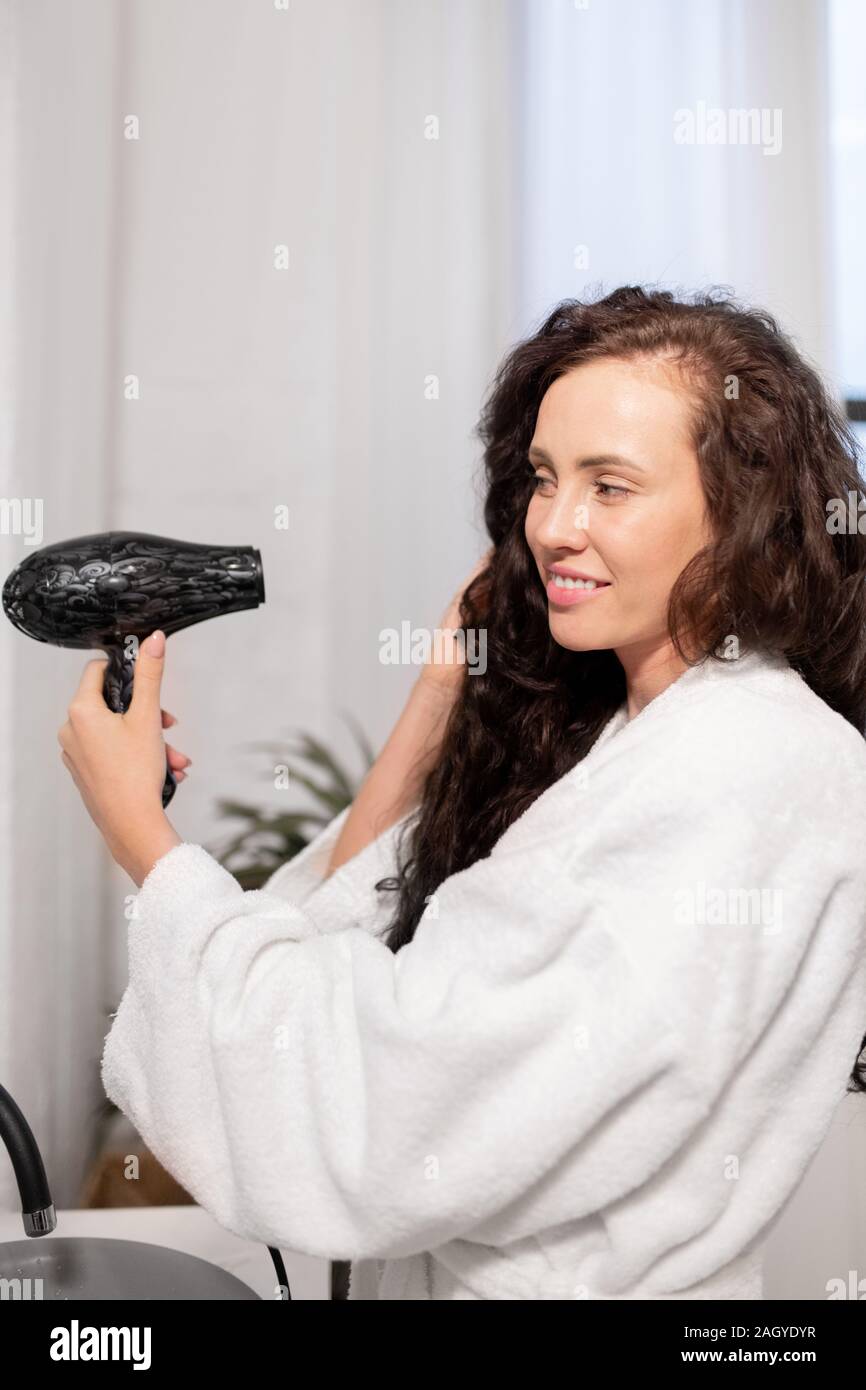 Smiling girl with hair-dryer taking care of her long wavy hair after washing Stock Photo