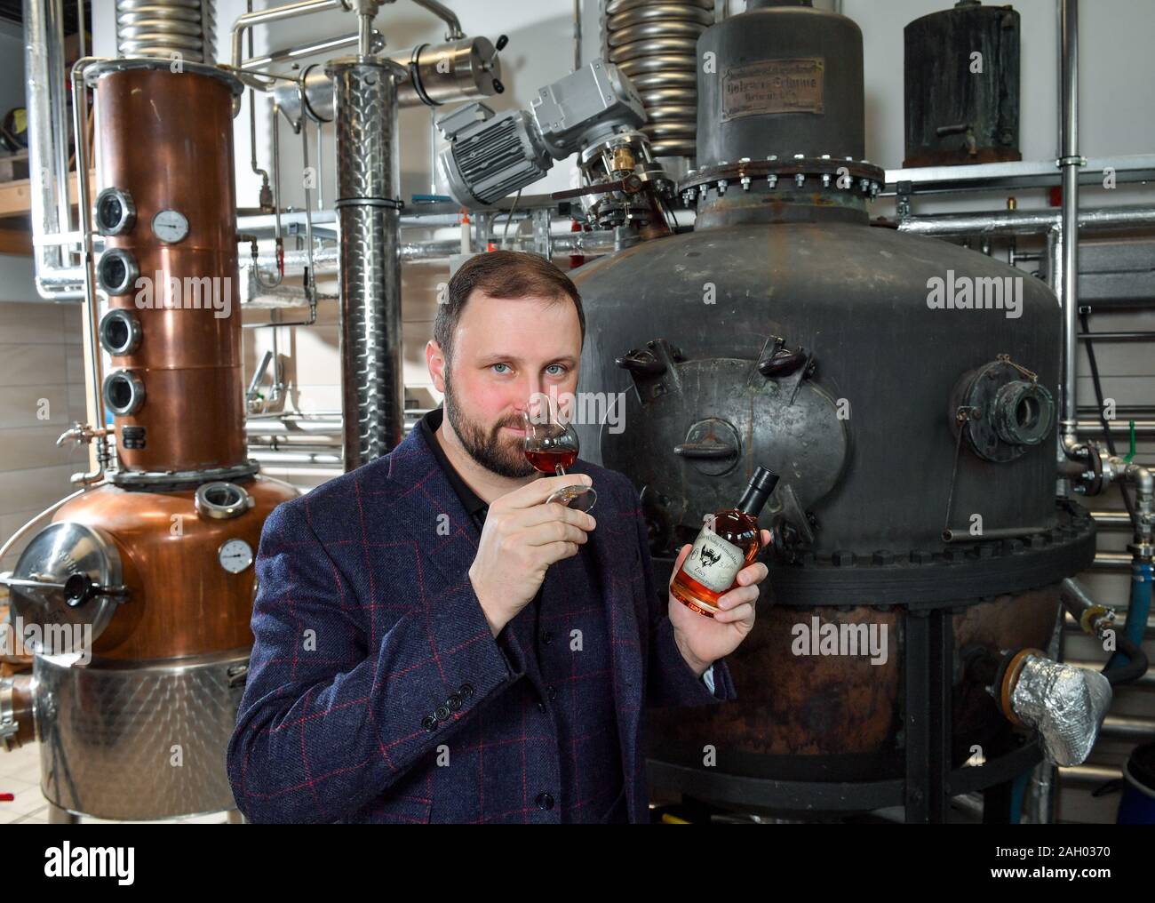 16 December 2019, Saxony-Anhalt, Zeitz: Daniel Rost, master distiller and owner of the Zeitzer Whisky Manufacture; tests a whisky. In the manufactory is the oldest whisky still in Germany (on the right in the picture) from 1935, from the mechanical engineering company Golzern-Grimma. Now the apparatus is the heart of the Zeitzer manufactory. It was first used in 1950 in Luckenwalde, Brandenburg, by the company C.W. Falckenthal Söhne. Since April, the historic still has been running again several times a month. About 22000 litres of pure alcohol burns rust per year in Zeitz. New barrels are reg Stock Photo