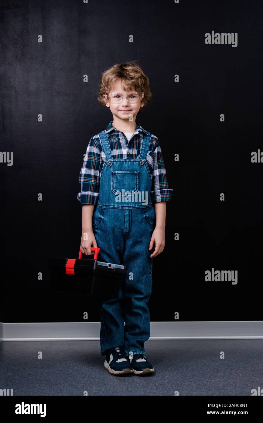 Happy little boy in denim overalls and eyeglasses holding toolbox while