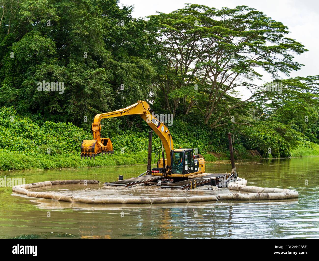 A yellow excavator / bulldozer performing dredging work in a canal / river in a scenic tropical rainforest / jungle habitat in Singapore, South-East A Stock Photo