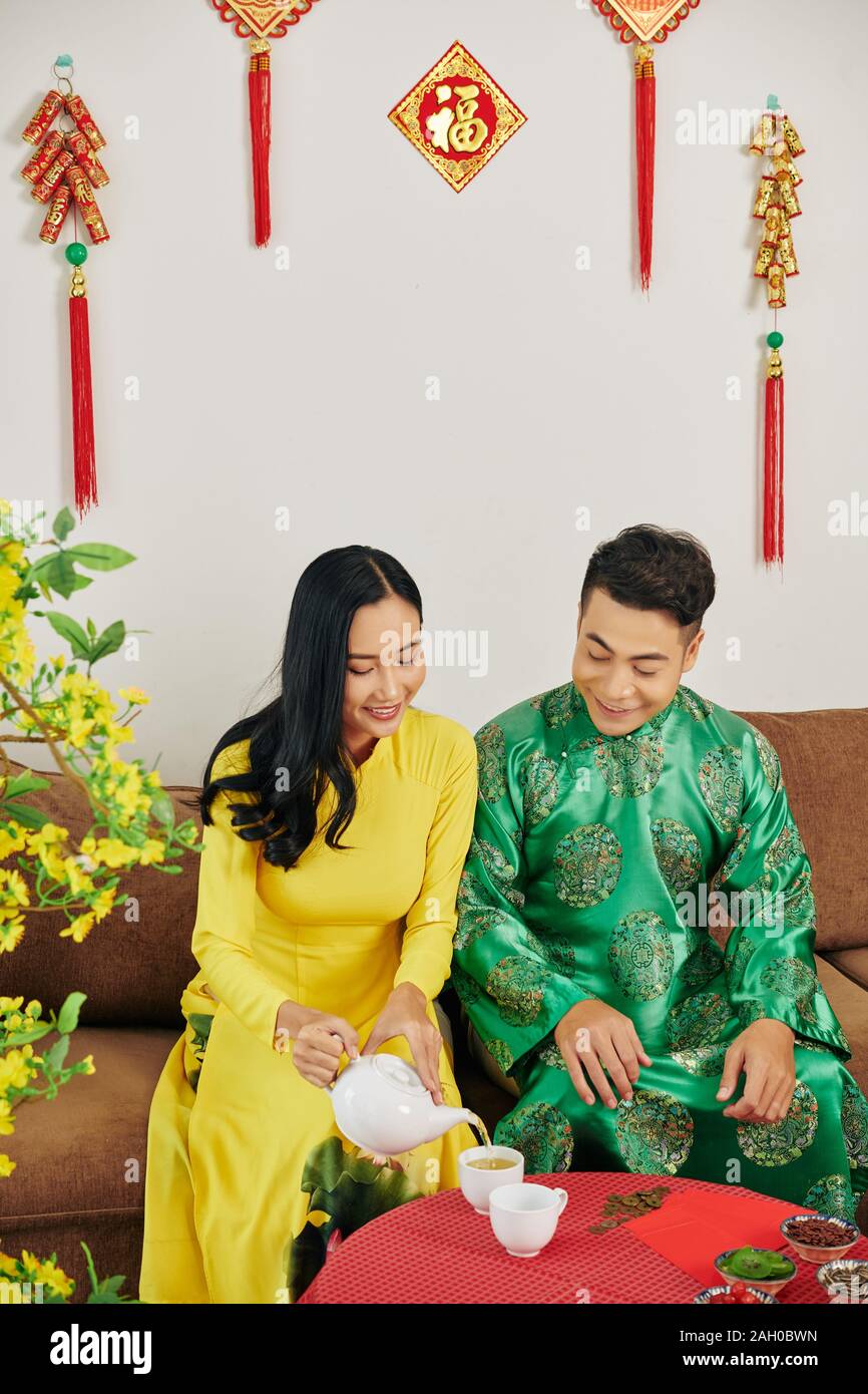 Young Vietnamese couple in traditionall dresses celebrating Lunar New Year and drinking herbal tea with dried berries Stock Photo