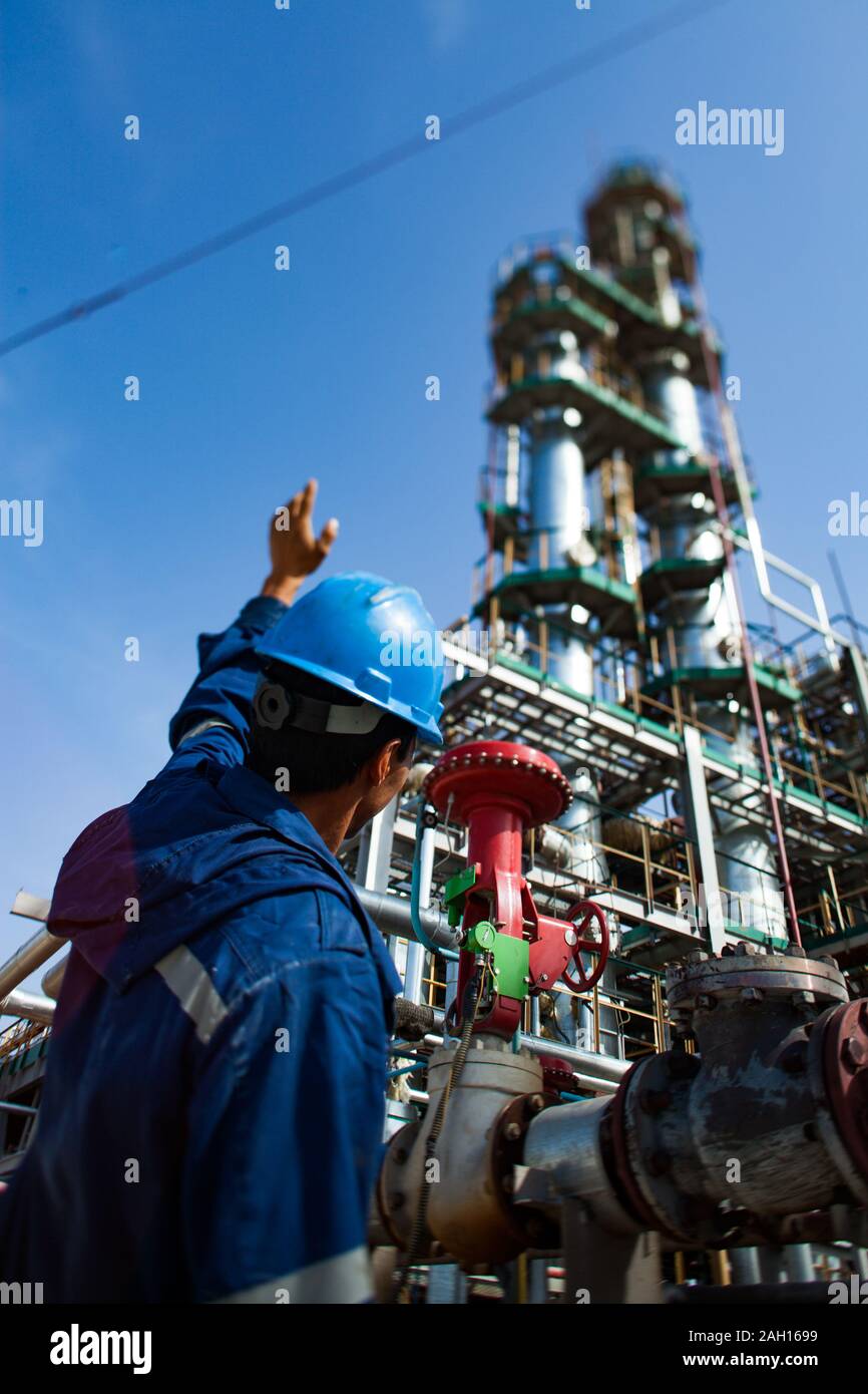 Worker in the blue uniform and blue helmet and grey distillation column at the oil refining plant on the blue sky. Stock Photo