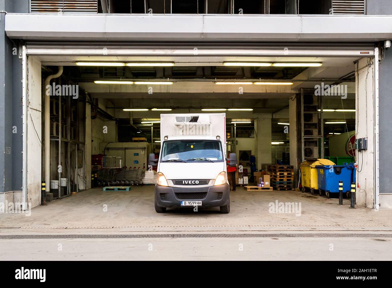 Varna, Bulgaria, December 20, 2019. White van while unloading goods in a delivery area with opened rolling gates behind a supermarket. Front view. Stock Photo