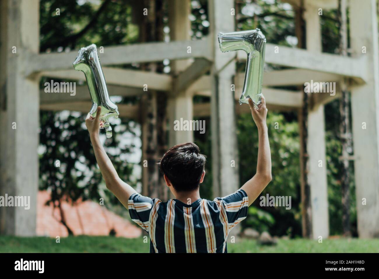 A young man facing away from the camera, wearing a blue-striped shirt, holding up silver balloons, '17', with one balloon in each hand. Stock Photo