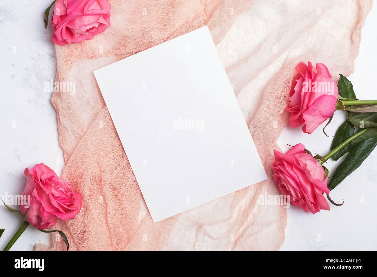 Top view of blank paper card lying on the gentle light pastel color cloth on the table with pink roses. Wedding or birthday template. Stock Photo