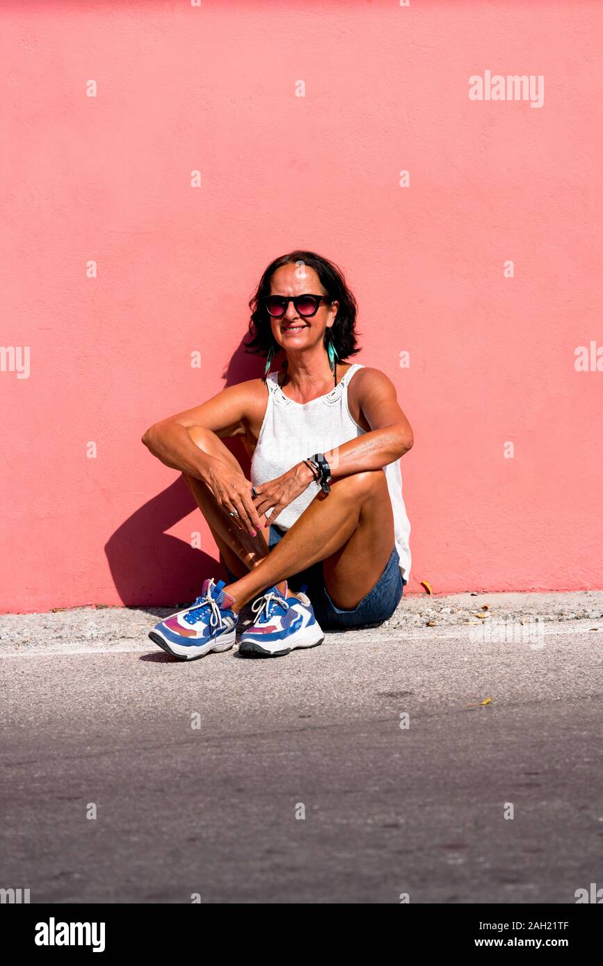 Attractive older woman with suntan wearing sunglasses leaning against a bright light pink monochrome wall. Sunny sunshine day one person older mature female. Stock Photo