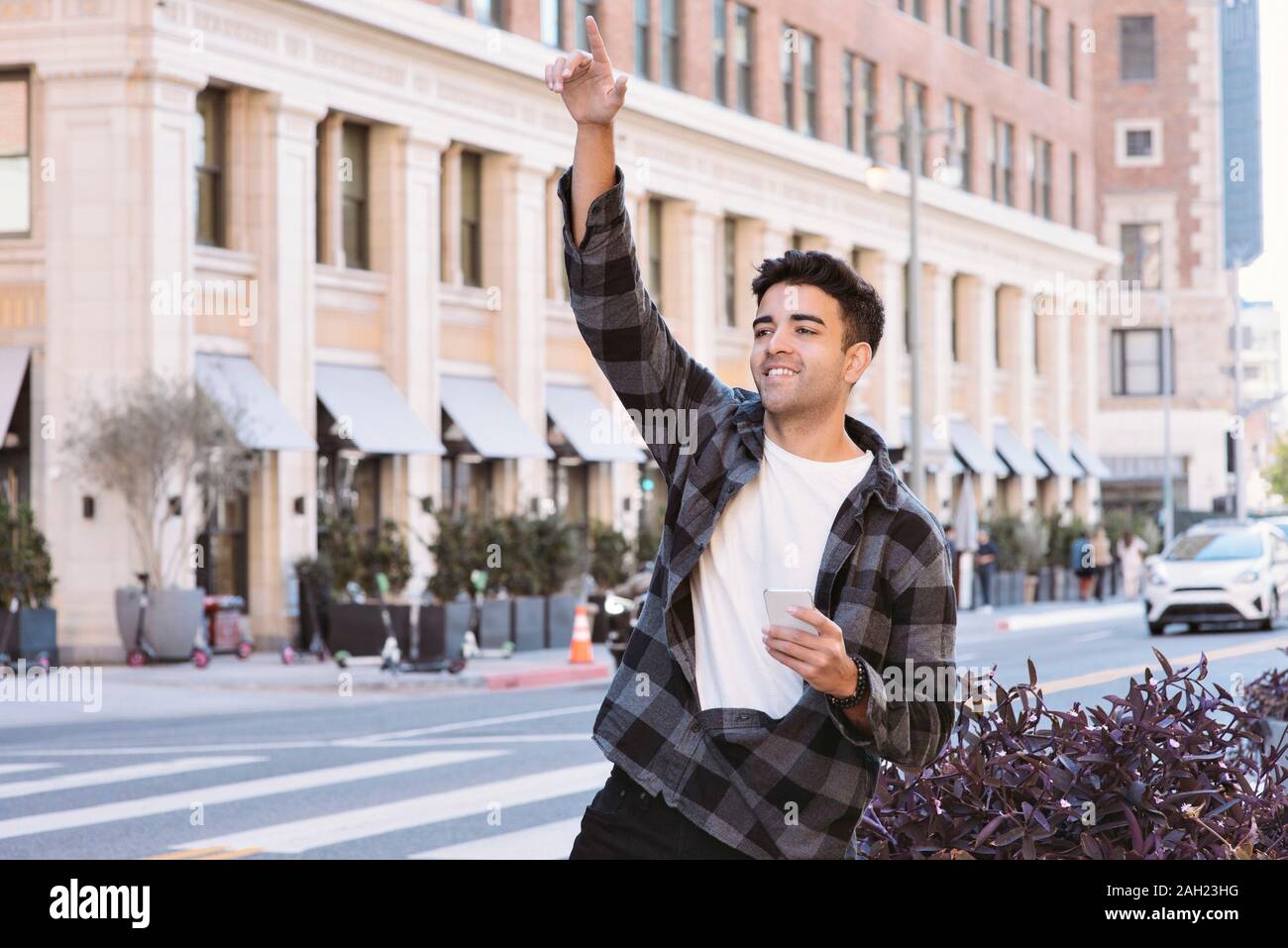 View of a Young Latin Man Hailing a Ride Share Car with a Cell Phone in the City - Afternoon with Bright Lighting Stock Photo