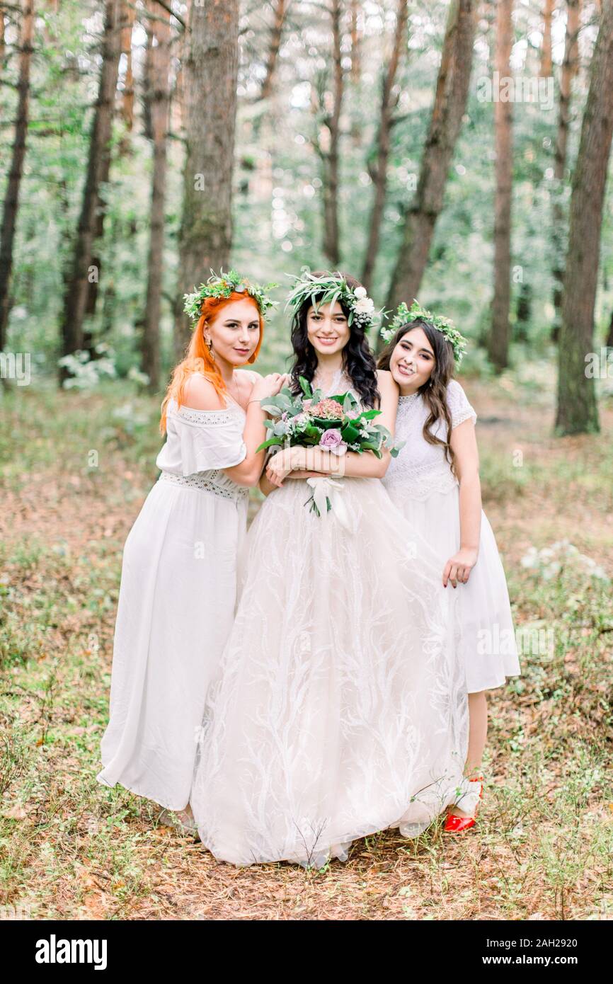 Rustic wedding outdoors. The bride in a white dress and floral wreath  standing with her two bridesmaids in green forest Stock Photo - Alamy