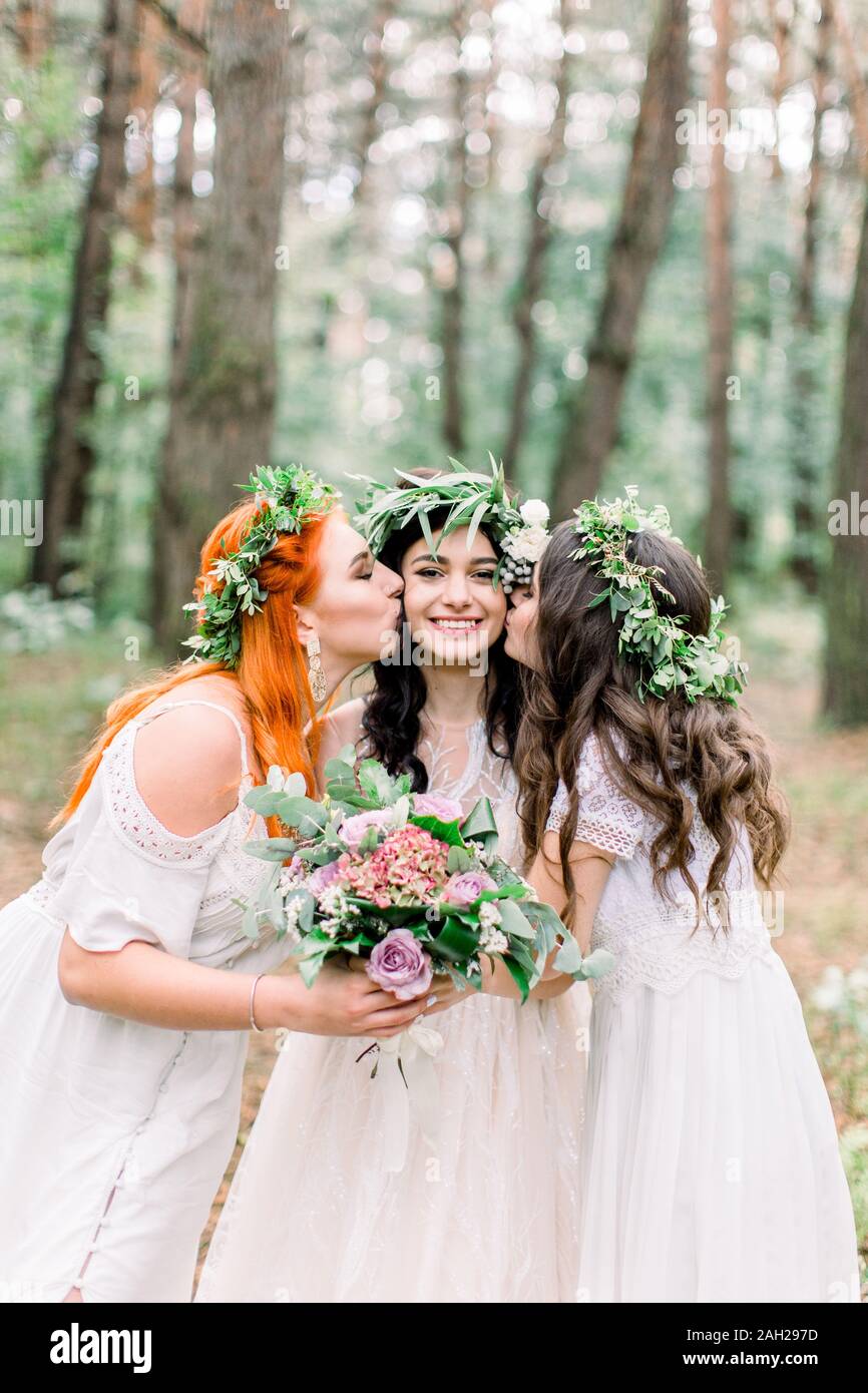 Two beautiful bridesmaids kiss bride's cheeks while they stand in the ...