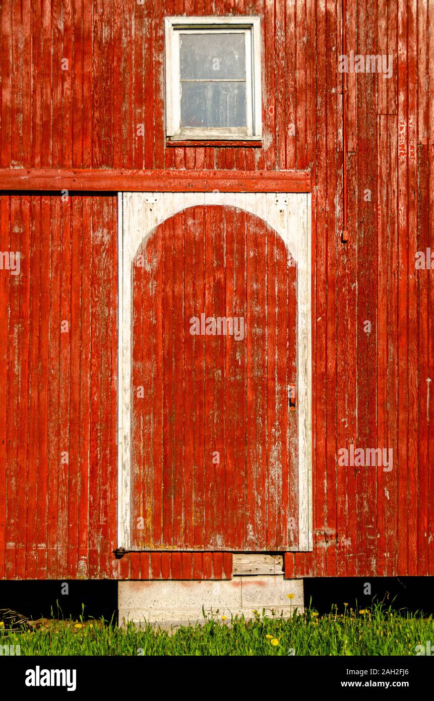 A closeup of a door and a window of red barn with weathered wooden exteriors illuminated by strong morning sunlight. Stock Photo