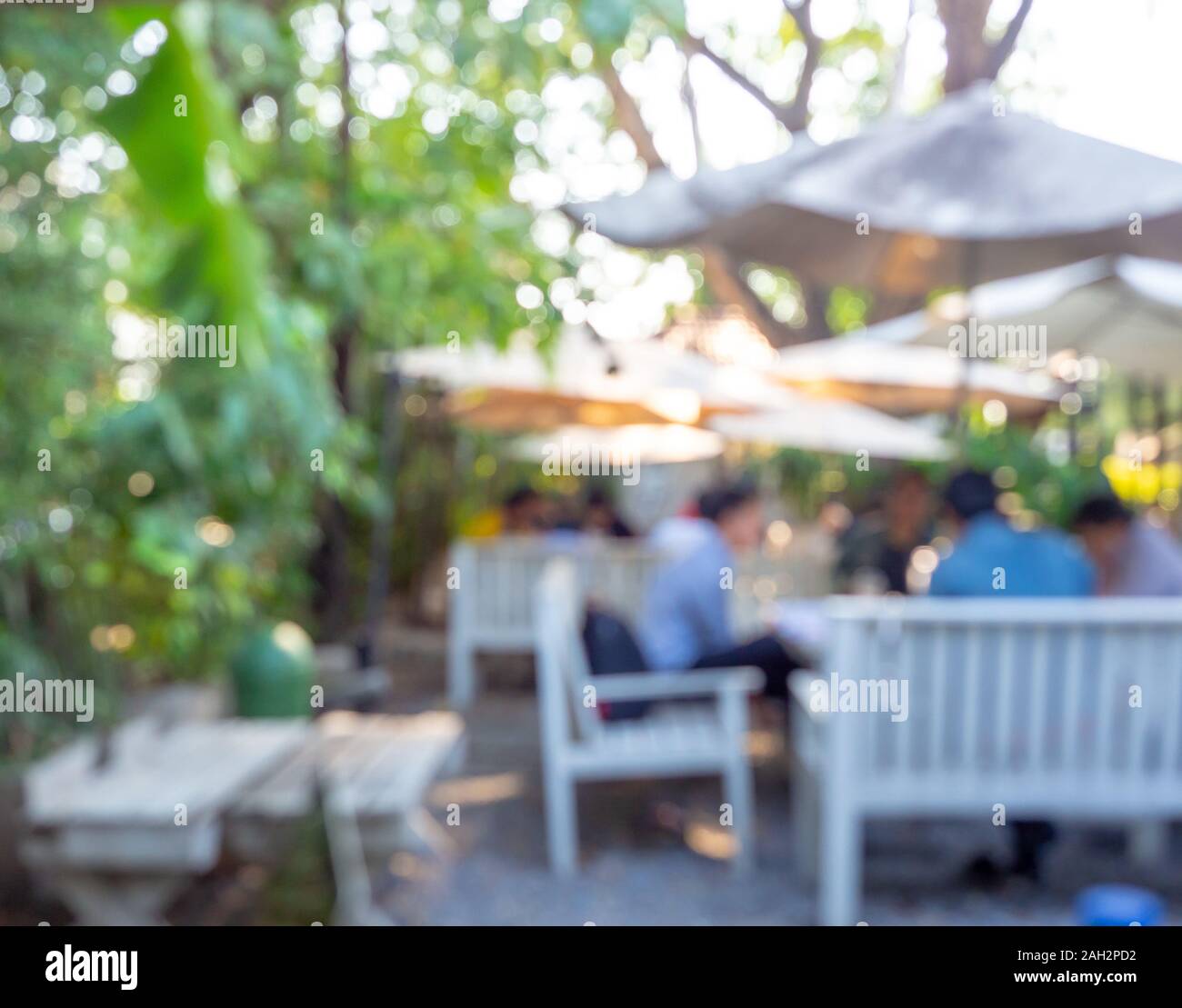 Blurred cafe outdoor background on sunny day. Blurry people sitting on white wooden chair under umbrella in the garden coffee shop with green tree. Stock Photo
