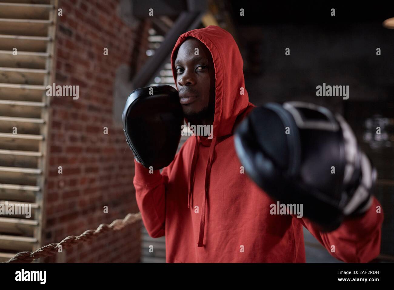 Portrait of young African boxer in hoody shirt wearing boxing gloves and posing at camera while standing in gym Stock Photo
