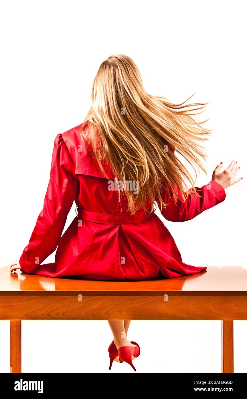 blond woman with long hair and wearing a red trench sitting on a table, view from the back Stock Photo