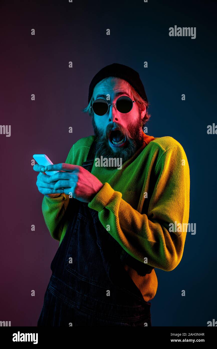 Using phone, extremely shocked. Caucasian man's portrait on gradient studio background in neon light. Beautiful male model with hipster style. Concept of human emotions, facial expression, sales, ad. Stock Photo