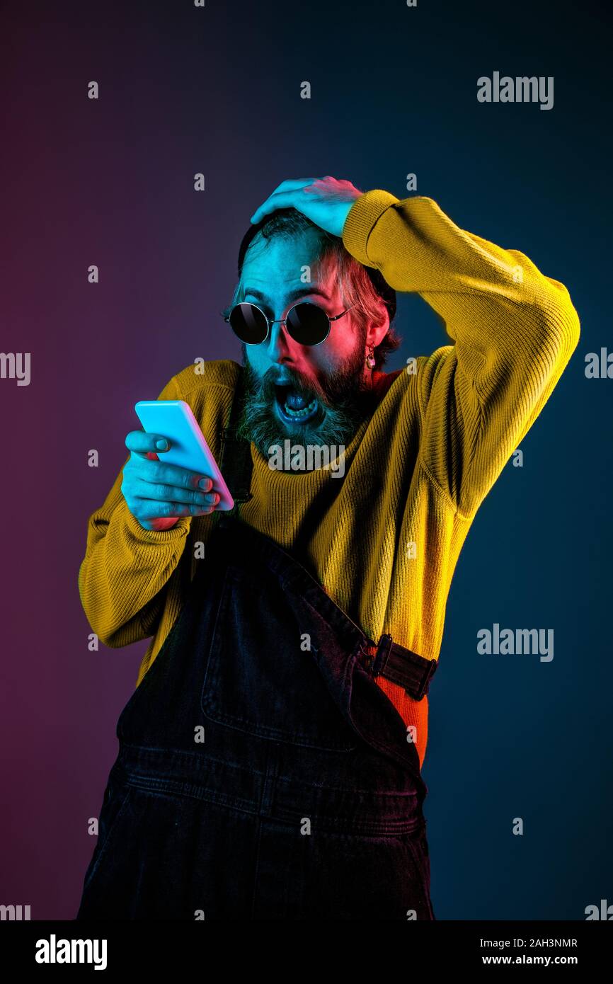 Using phone, extremely shocked. Caucasian man's portrait on gradient studio background in neon light. Beautiful male model with hipster style. Concept of human emotions, facial expression, sales, ad. Stock Photo