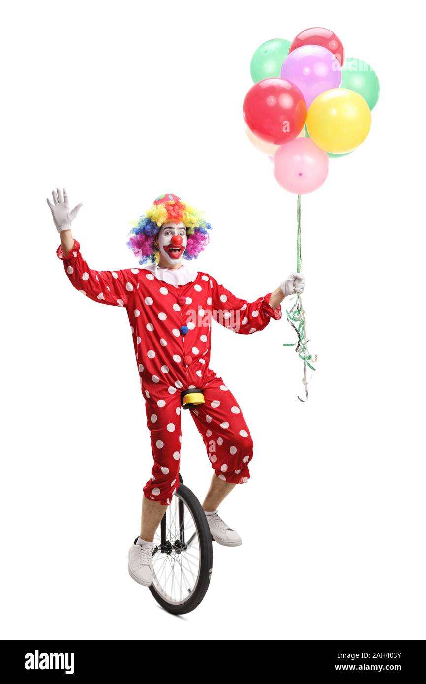 Happy clown riding a unicycle, holding a bunch of balloons and waving at the camera isolated on white background Stock Photo