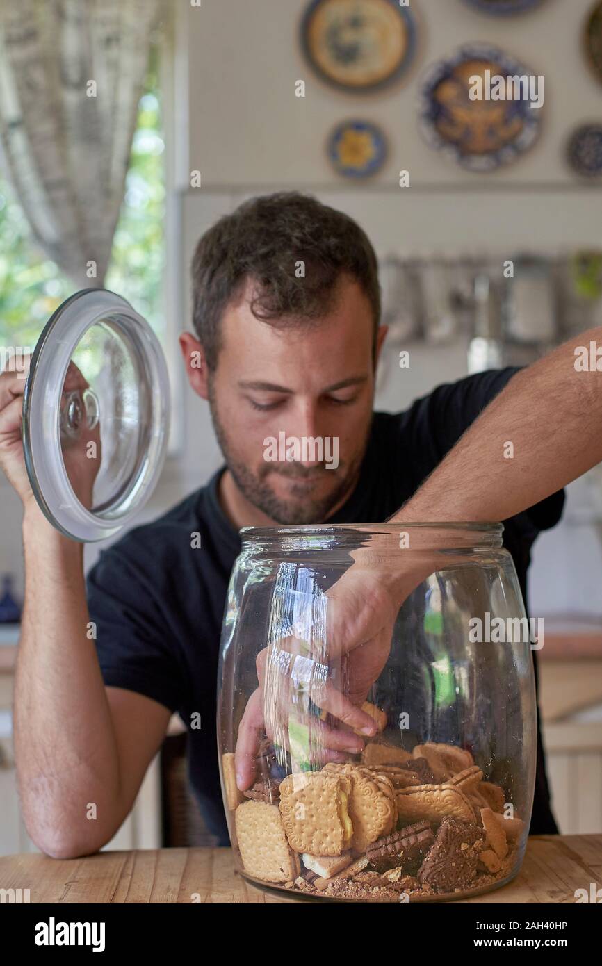 Man sitting in kitchen, looking in cookie jar, taking cokkie Stock Photo