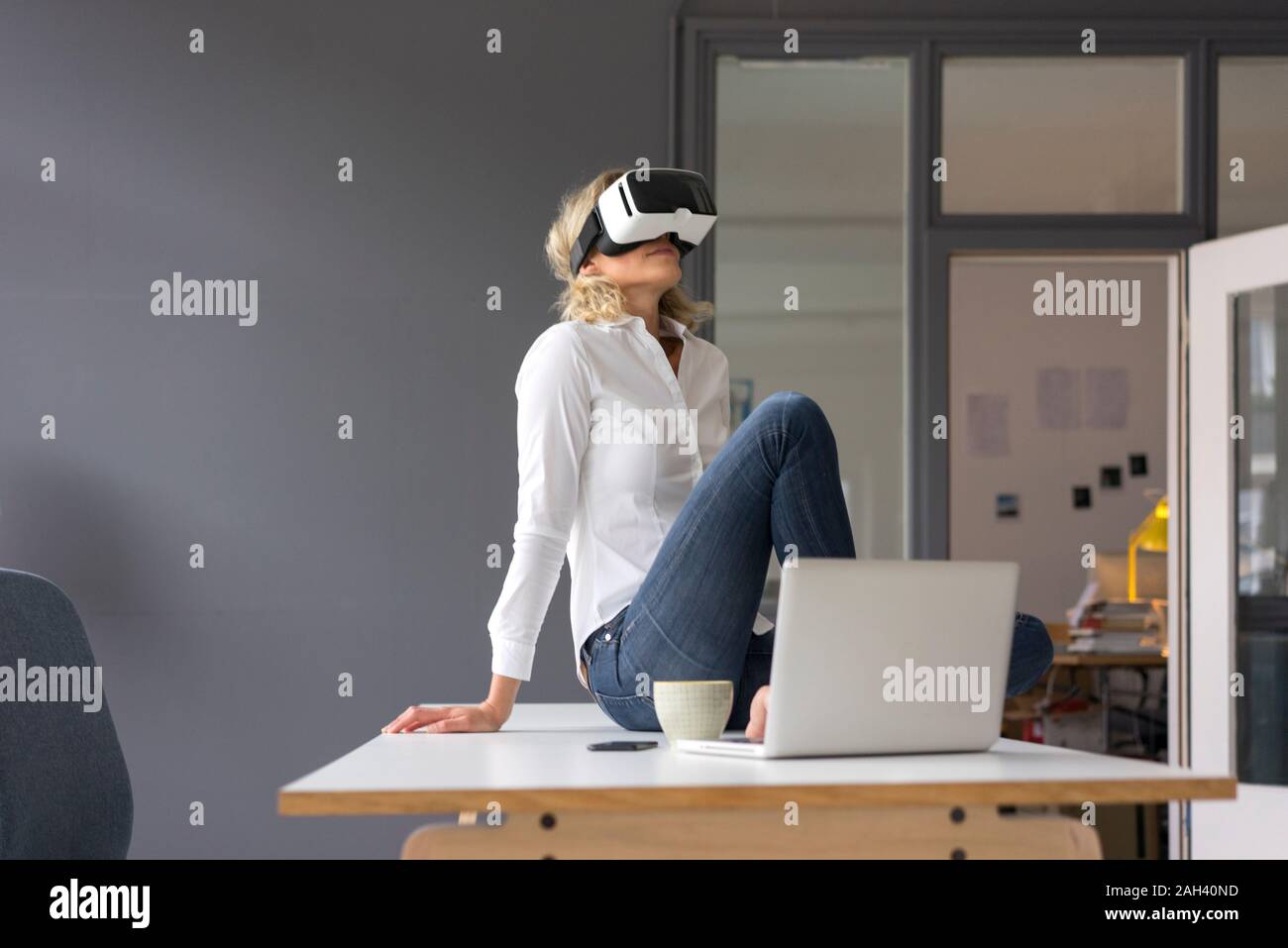 Young businesswoman sitting on desk in office wearing VR glasses Stock Photo
