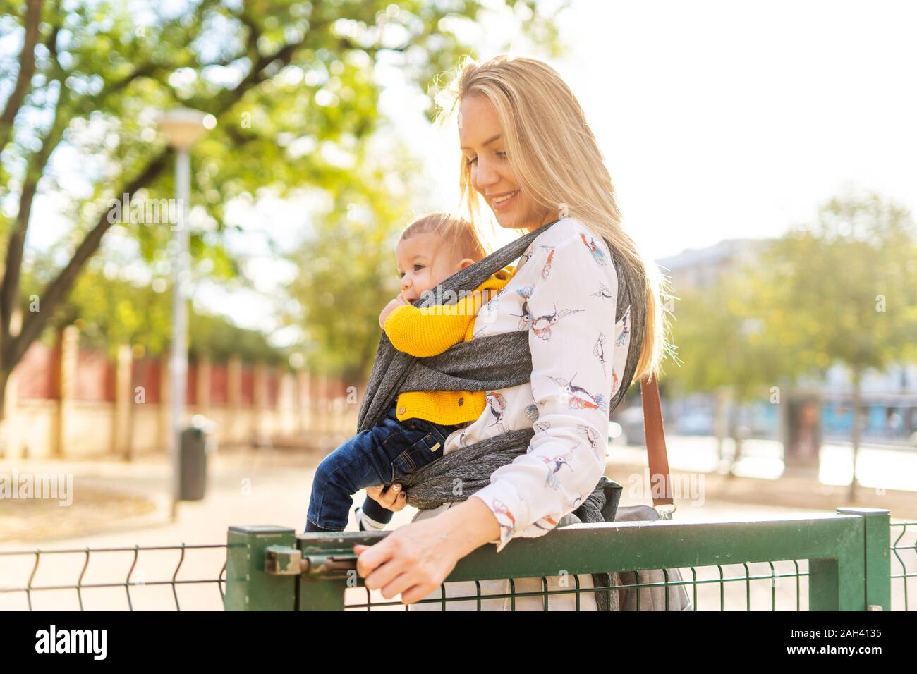 Mother carrying baby boy in a sling opening a gate Stock Photo