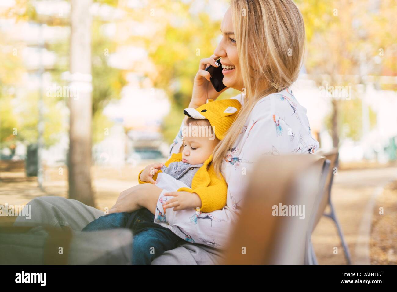 Happy mother resting with baby boy on a park bench talking on the phone ...
