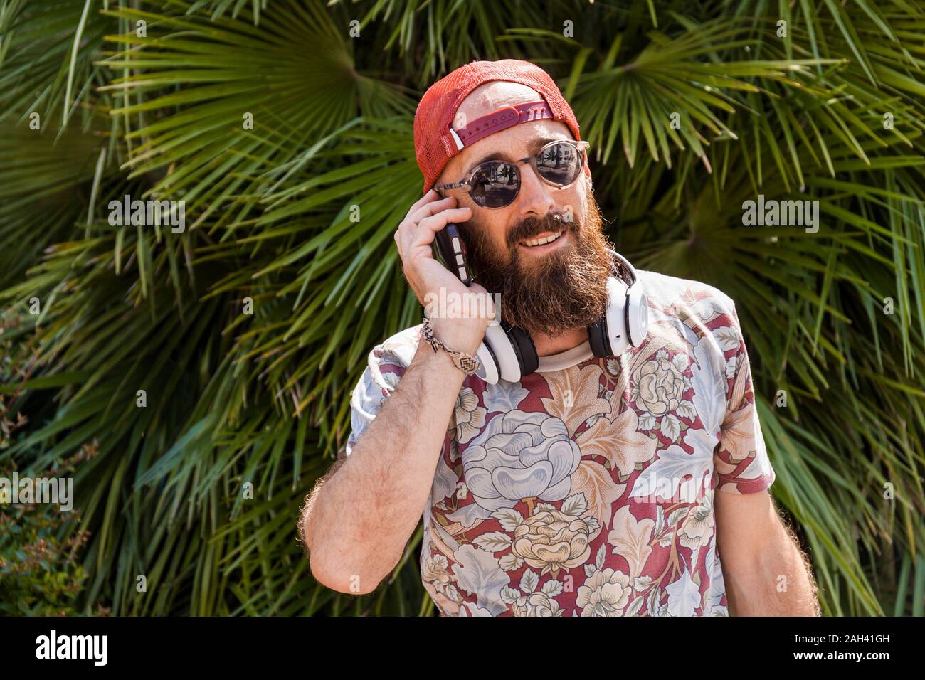 Mature man with red basecap, sunglasses and white headphones using smartphone Stock Photo