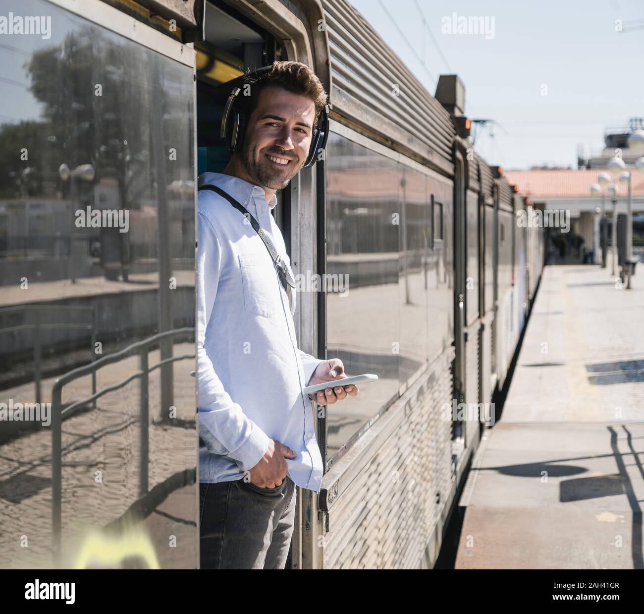 Smiling young man with headphones and smartphone standing in train door Stock Photo