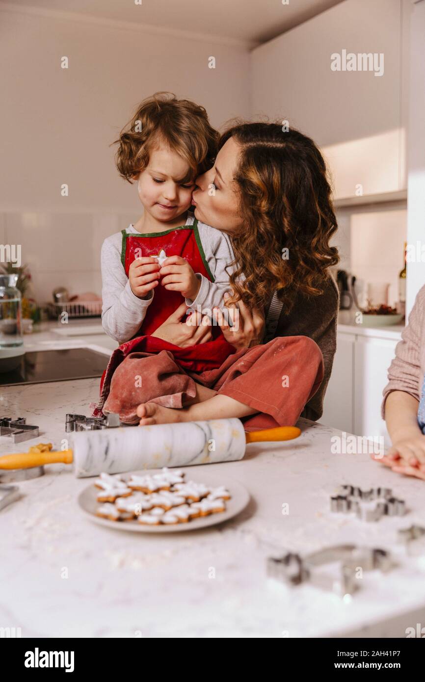 Mother kissing daughter in kitchen with Christmas cookies on counter Stock Photo