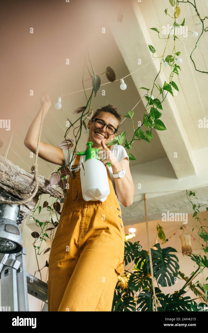 Happy young woman standing on a ladder caring for plants in a small shop Stock Photo