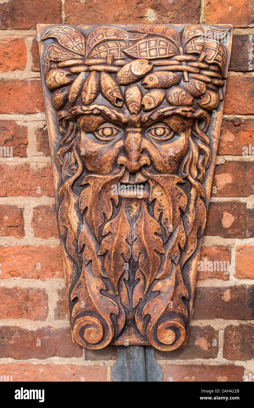 Ornate green man head and face sculpture with unusual crown of fishes hanging on red brick wall, Green Man Ceramics by Paul Gibson Stock Photo