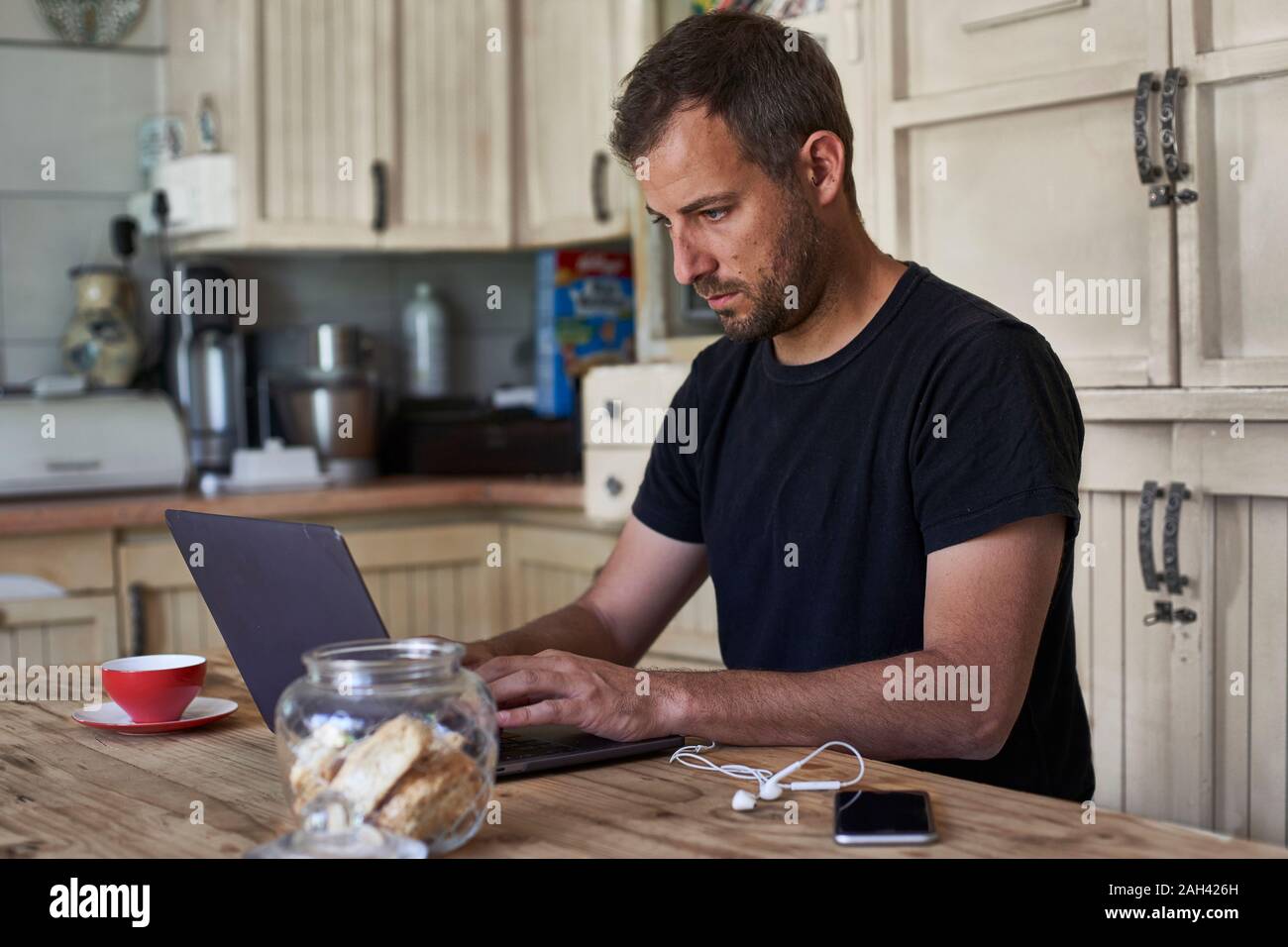 Man working from home, sitting at kitchen table, using laptop and smartphone Stock Photo