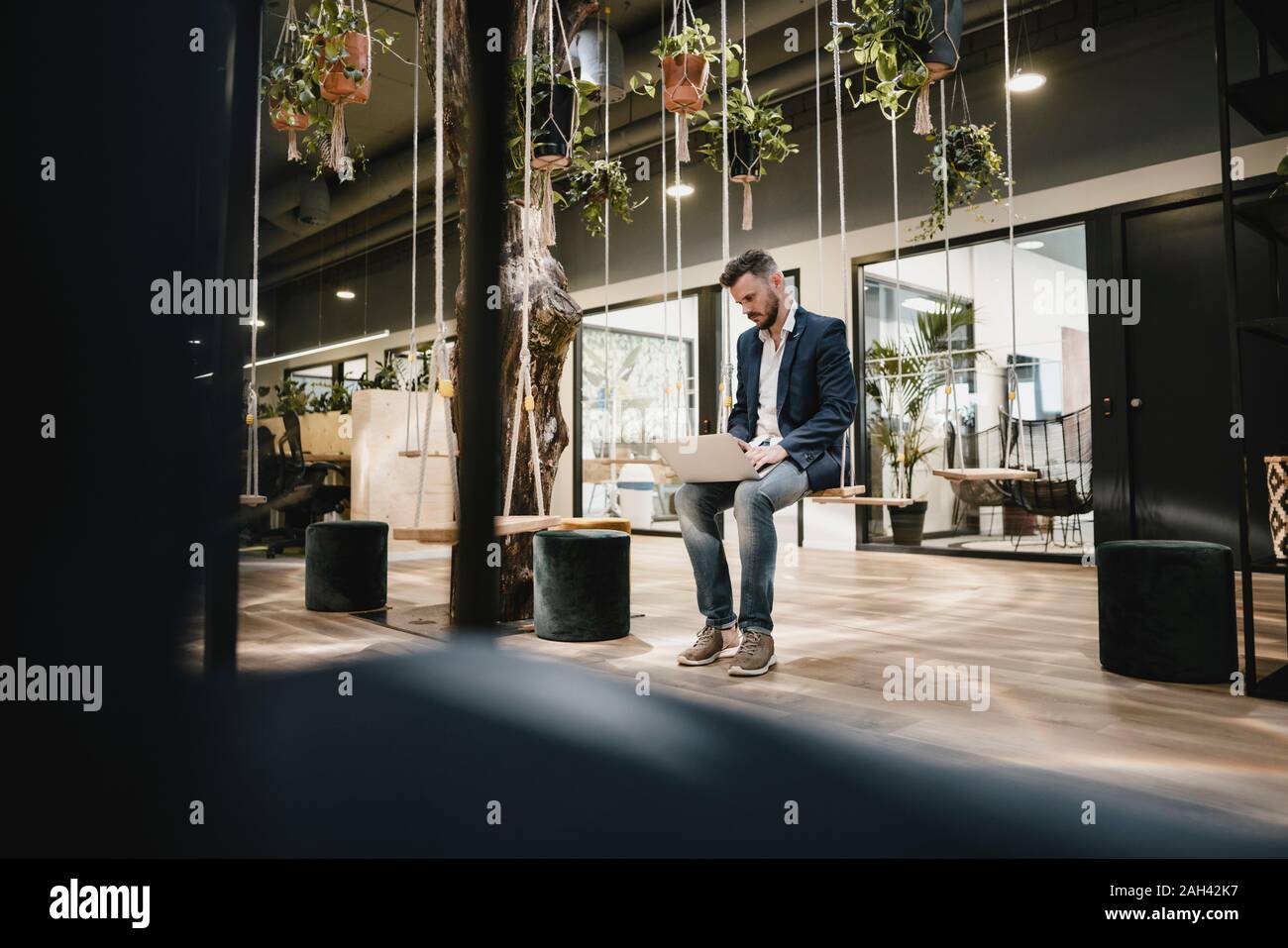 Businessman using laptop in coworking space, sitting on swing Stock Photo