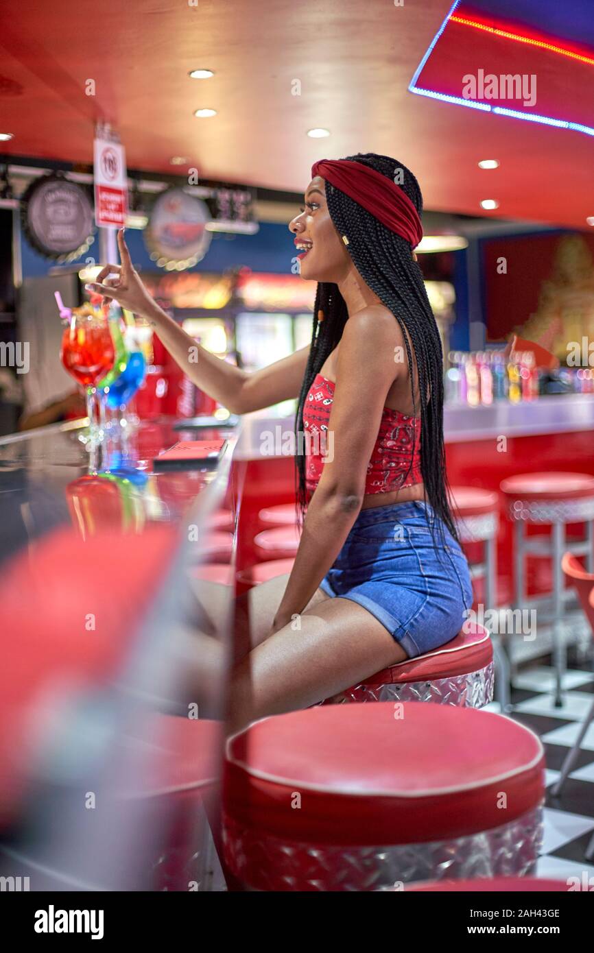 Young black woman with braided hairstyle sitting on a restaurant's bar red stool and ordering a drink Stock Photo