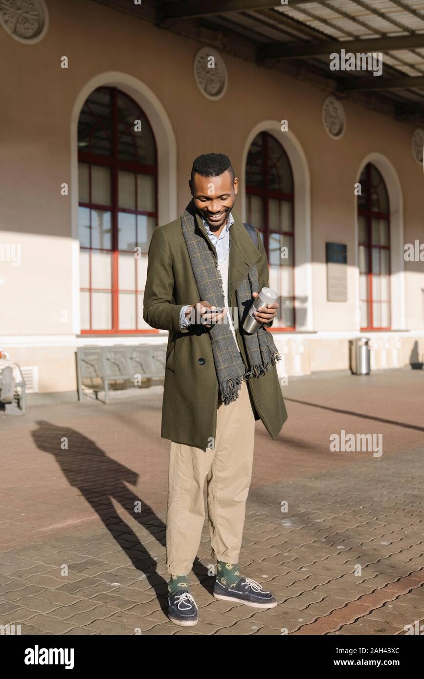 Stylish man with reusable cup using smartphone while waiting for the train Stock Photo