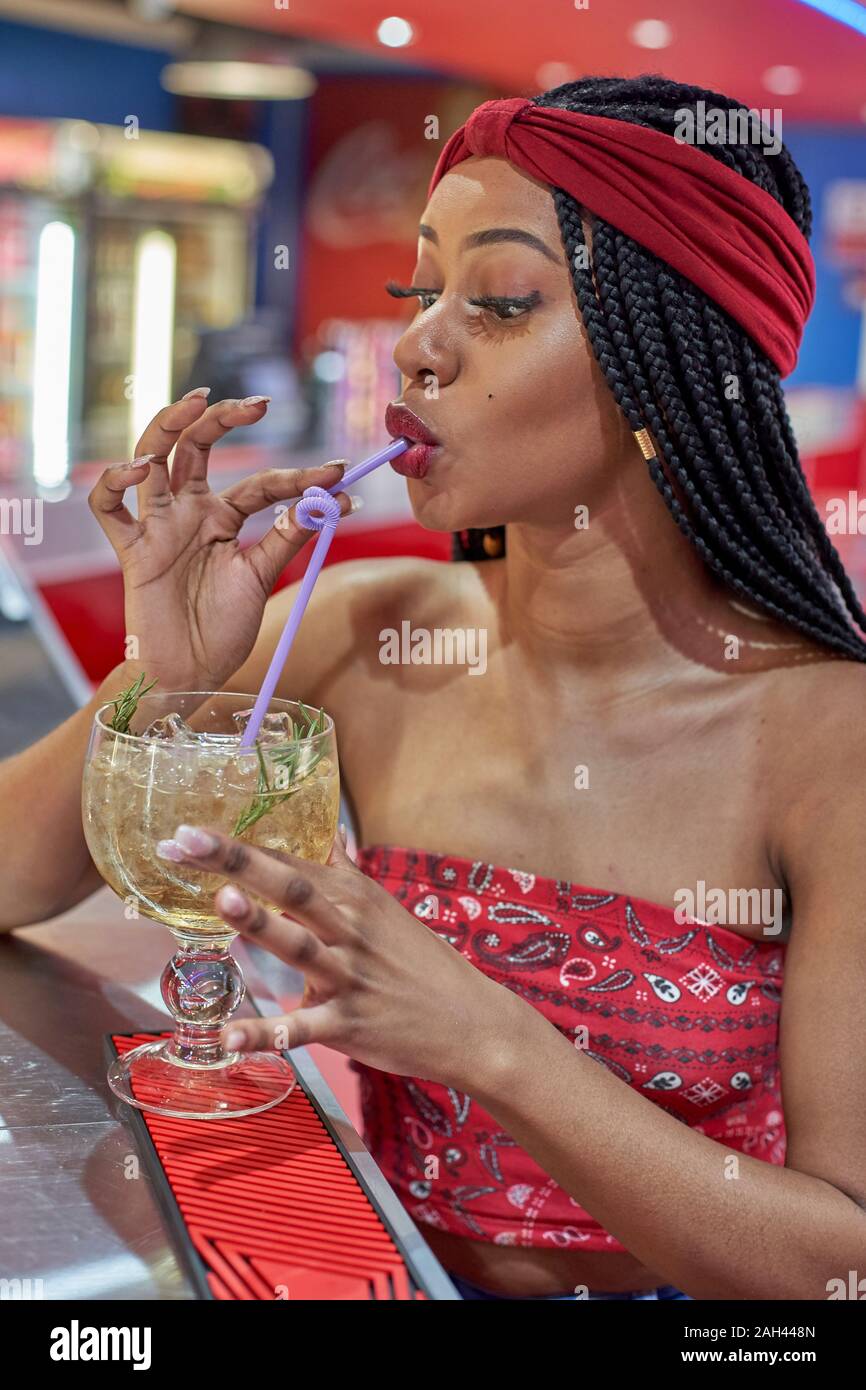 Young woman with braided hairstyle sitting on a restaurant's bar, drinking a cocktail with a straw Stock Photo