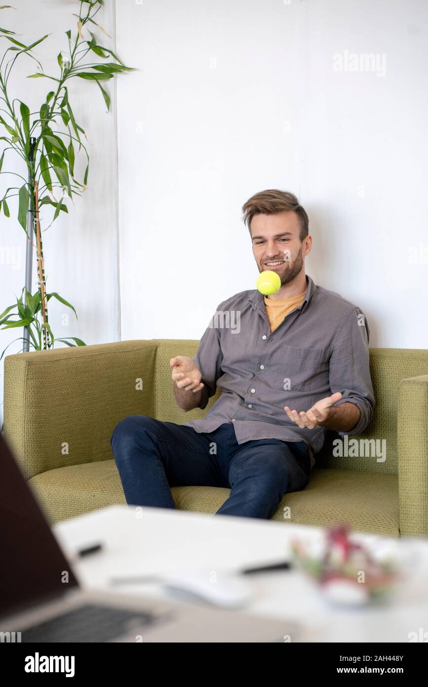 Smiling man sitting on couch in office playing with a tennis ball Stock Photo