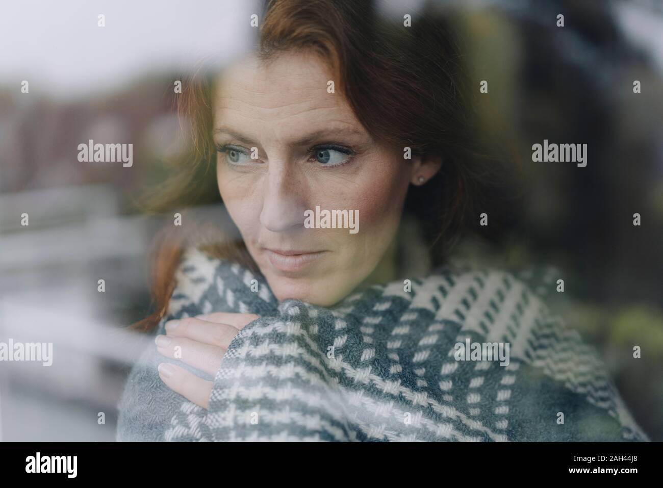 Woman looking out of window, wrapped in blanket Stock Photo